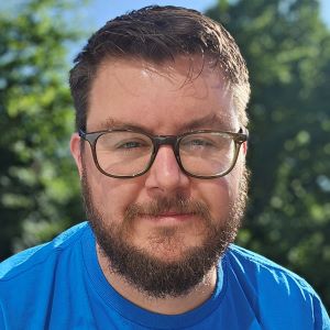 Ben has short dark hair and a matching beard. He is wearing dark rimmed glasses and a royal blue t-shirt. It's a sunny day, and Ben has a relaxed look while facing the camera.