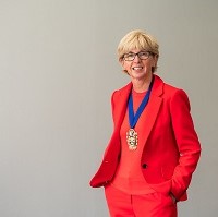 Caroline is wearing a red trouser suit, a red crew neck top, her gold coloured CIPFA president's medal which is hung from a blue ribbon. She has short blonde hair, dark rimmed glasses, is facing the camera while smiling and is set against a light grey background.