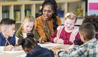 Children sat around a desk writing on pieces of paper with their teacher in the middle